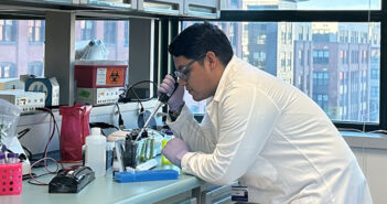 Christian Godinez leans over a counter while performing research in a lab with the Providence skyline visible outside of the window.