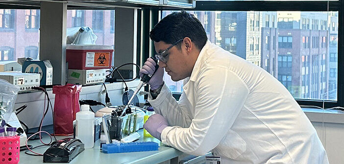 Christian Godinez leans over a counter while performing research in a lab with the Providence skyline visible outside of the window.