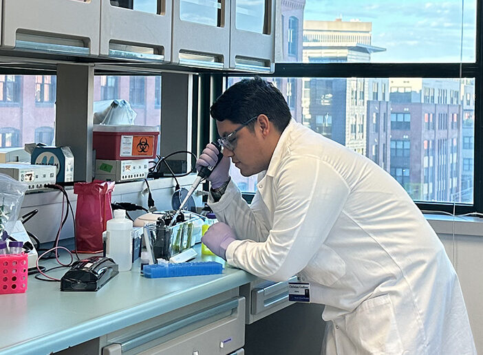 Christian Godinez leans over a counter while performing research in a lab with the Providence skyline visible outside of the window.